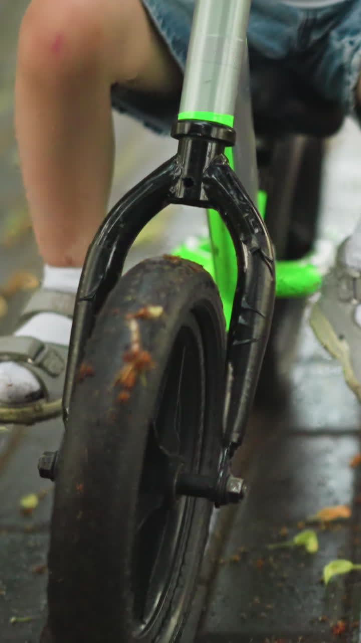 primer plano de un niño en una bicicleta con una mujer caminando cerca de él en un camino cubierto de hojas, el niño deja caer su pierna para el equilibrio mientras maniobra a través del camino