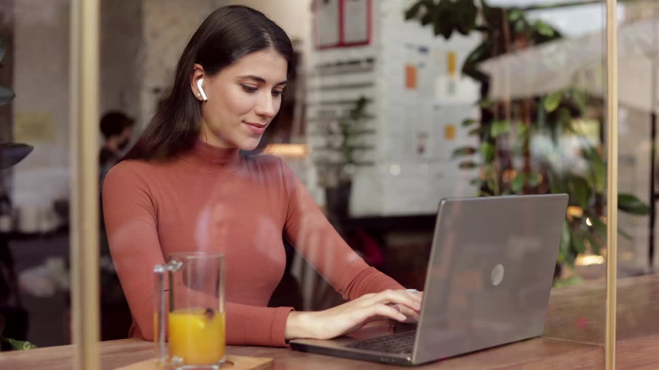 mujer freelance sonriente trabajando en una computadora portátil sentada en una cafetería. escuchando música usando inalámbrico. mensajes de texto, comunicación en línea con colegas, parientes, amigos. trabajo remoto