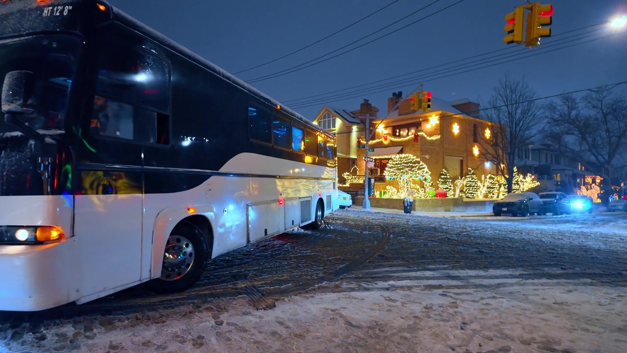 Coach bus at snowy Dyker Heights Christmas display in Brooklyn. Night view shows an idling bus, traffic signals and holiday lights outside residential homes.
