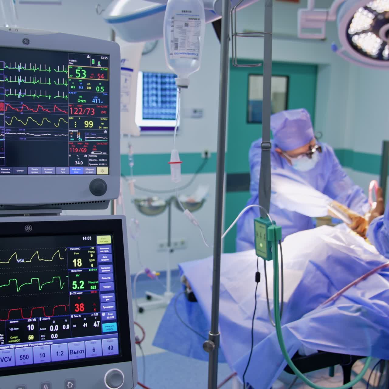 Zwei Monitore mit Lebensparametern des Patienten. NeurochirurgInnen arbeiten am Operationstisch im Hintergrund