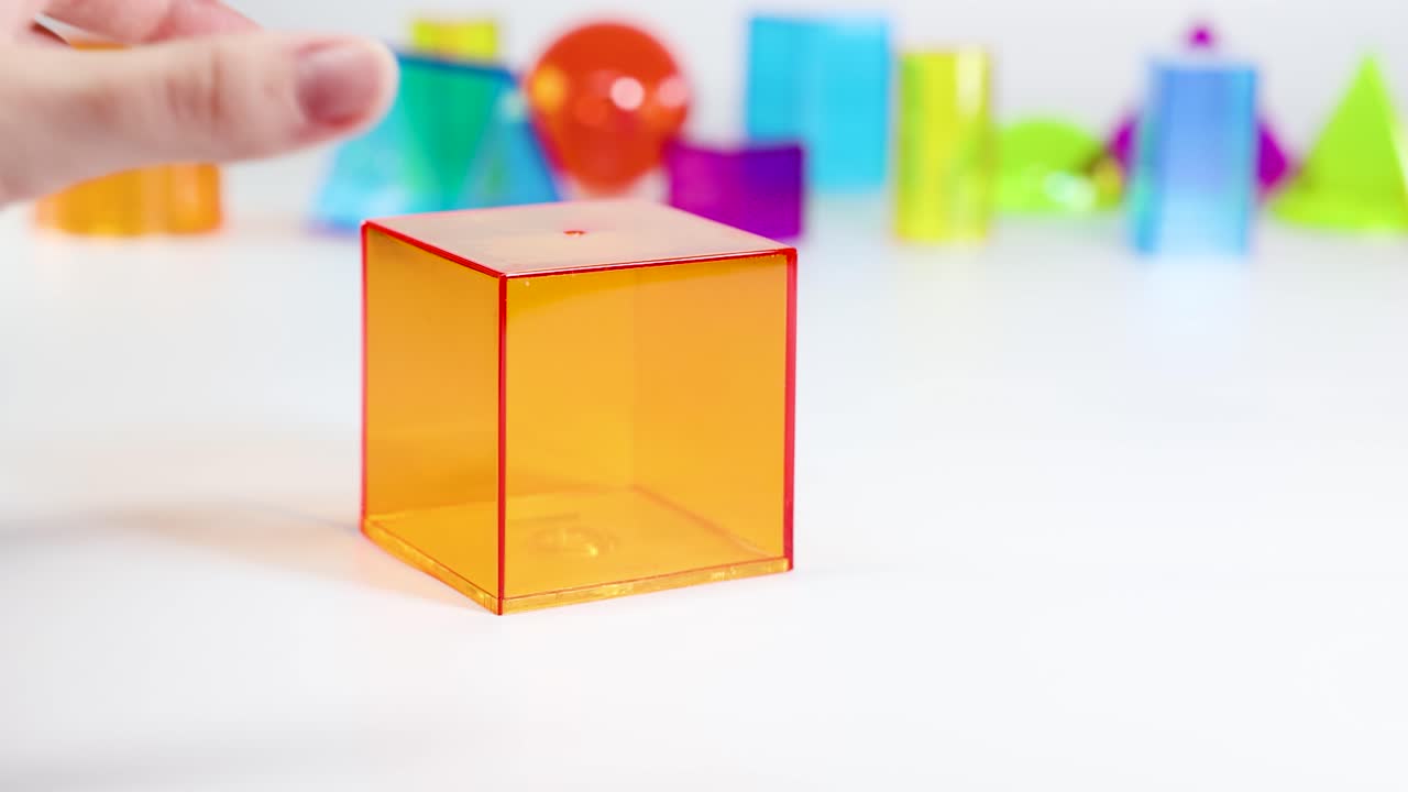 A hand manipulates a transparent orange cube among various colorful geometric shapes on a white surface