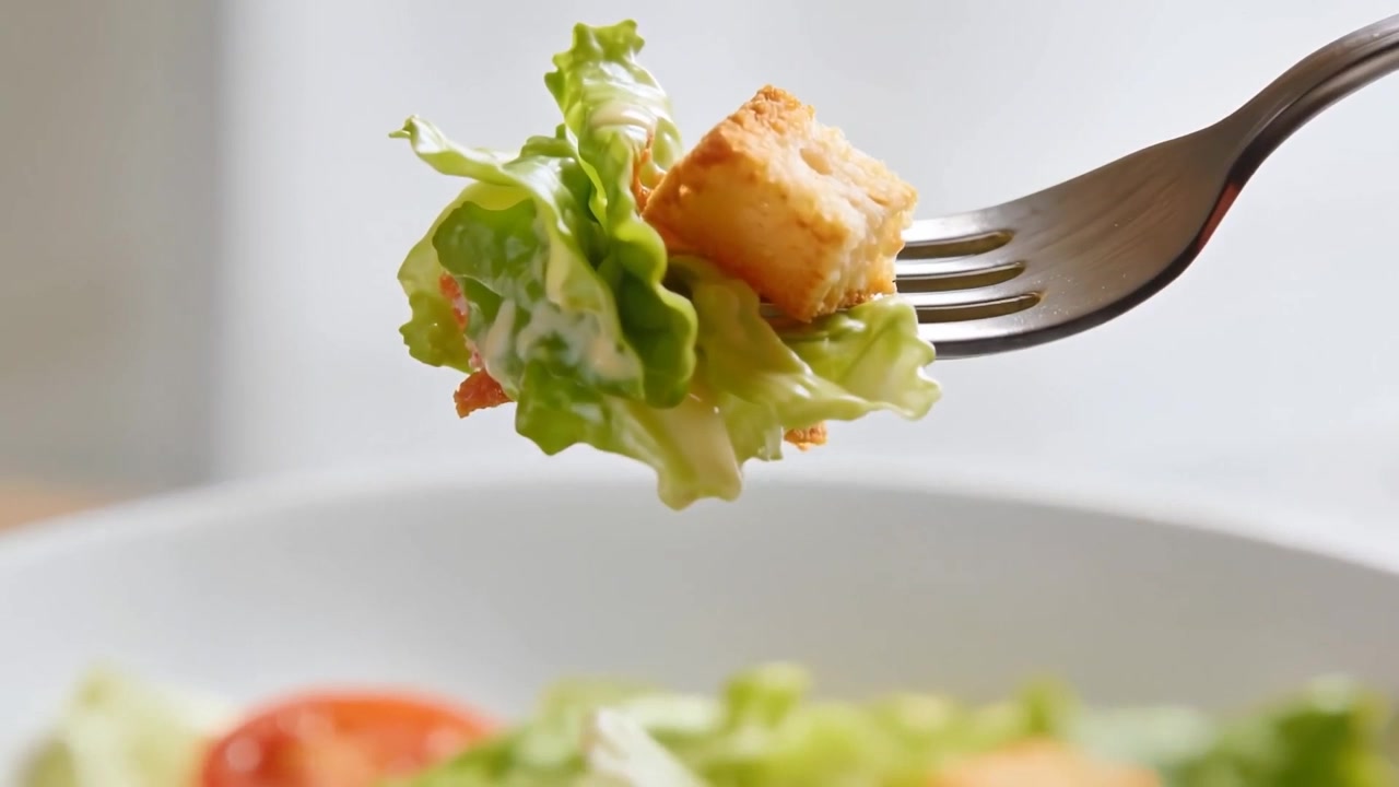 Lifting metal fork entering right, carrying lettuce and crouton with dressing at table for closeup