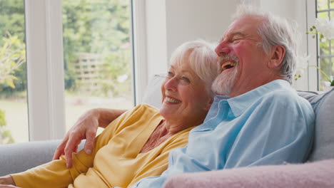 Pareja-De-Ancianos-Jubilados-Con-Control-Remoto-Sentados-En-Un-Sofá-En-Casa-Viendo-Televisión-Y-Riendo-Juntos