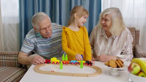 Child-girl-kid-involved-in-build-board-game-ride-toy-train-on-railroad-with-senior-grandparents-home