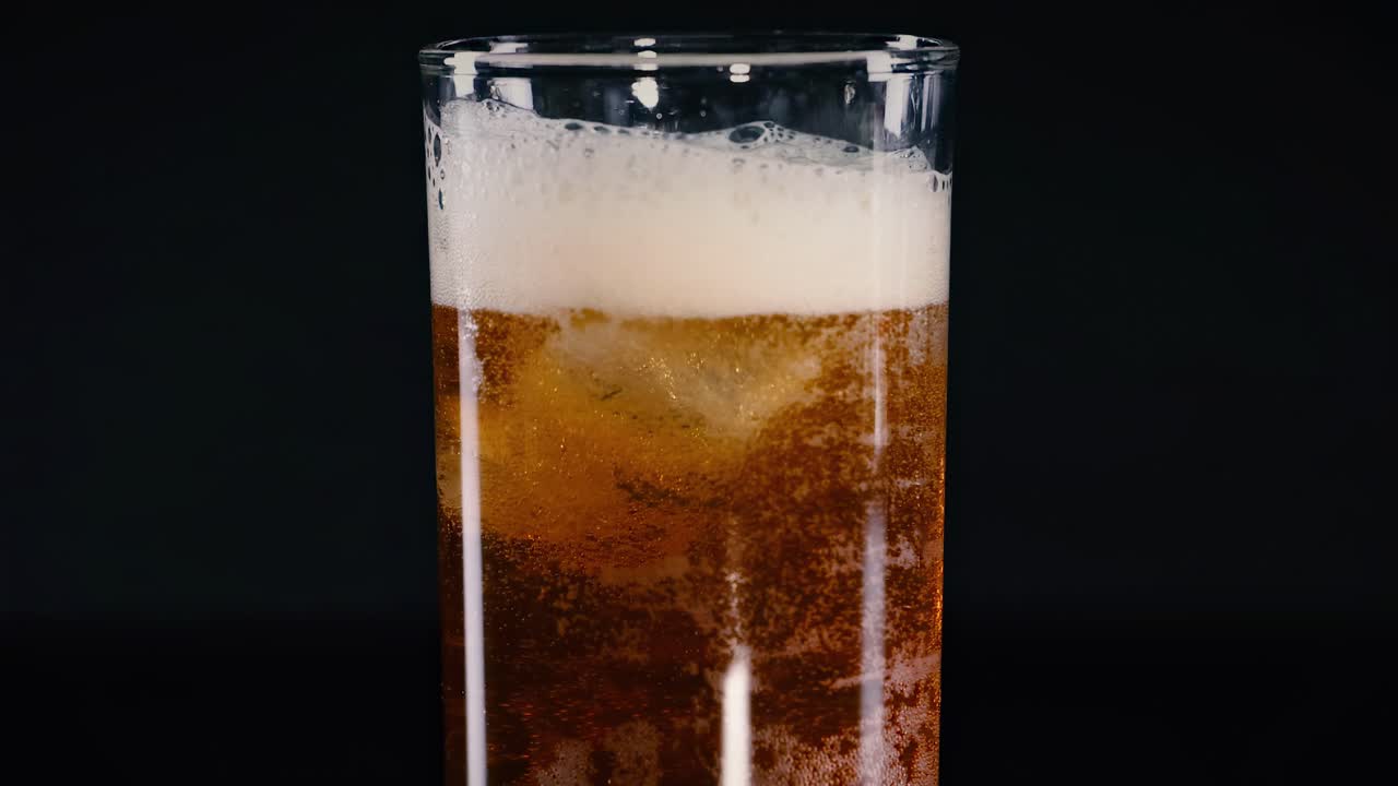 A tall glass filled with a foamy amber beverage against a dark background, showcasing bubbles and ice.