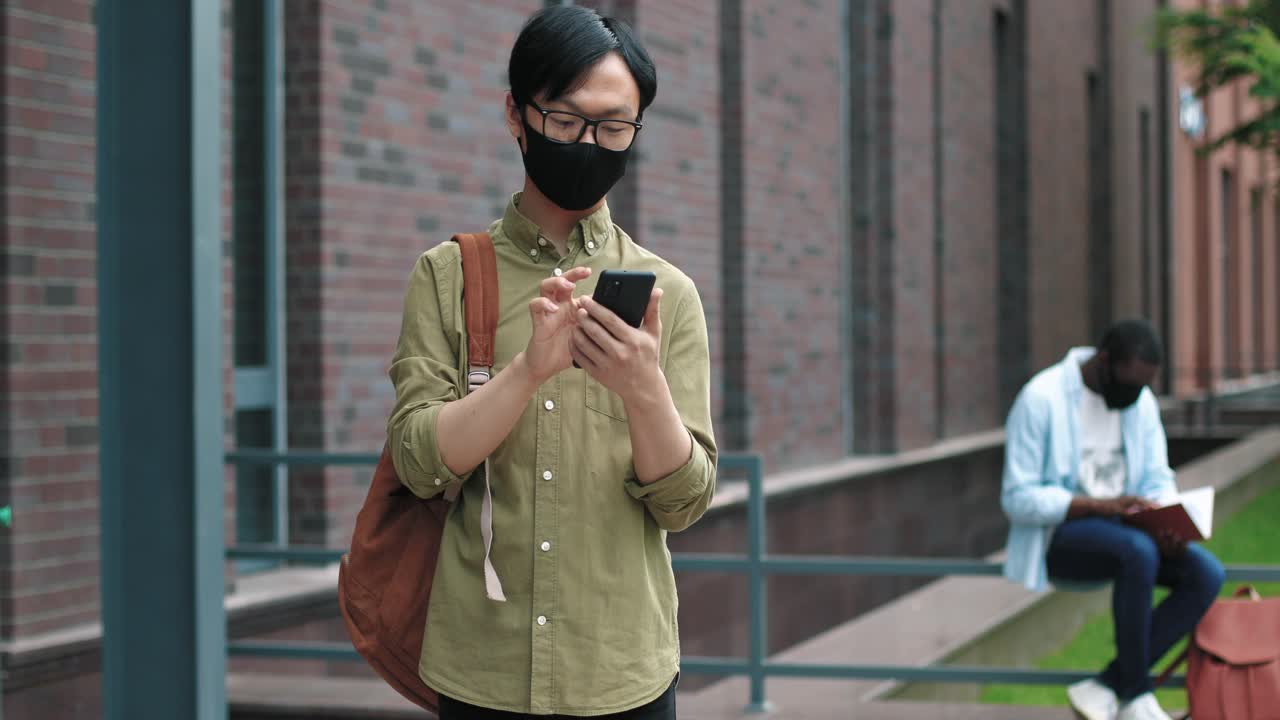 Asian student wearing glasses and facial mask holding his smartphone near the college