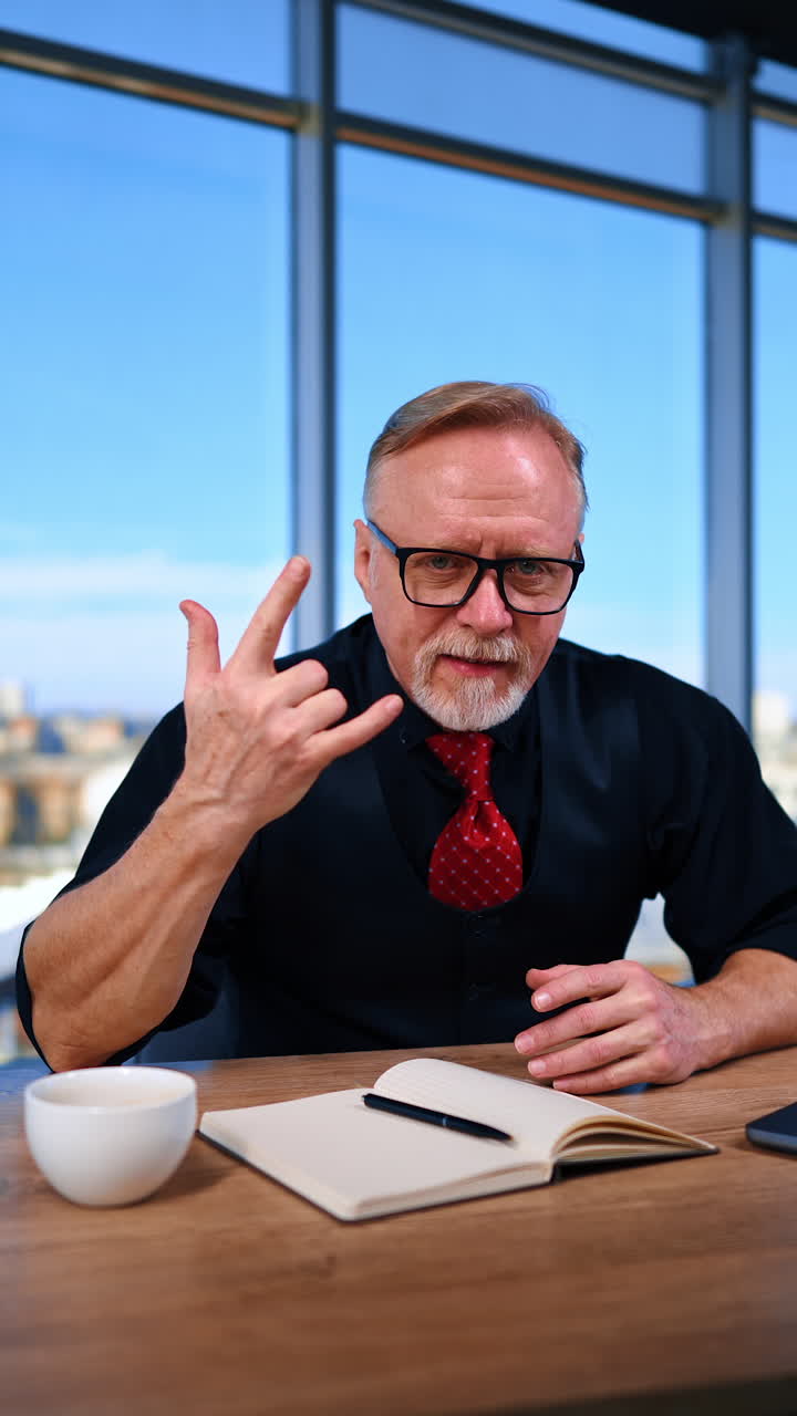 Mature bearded Caucasian male in black clothes and red tie sits at desk. Man looks at camera showing rock hand gesture. Vertical video.