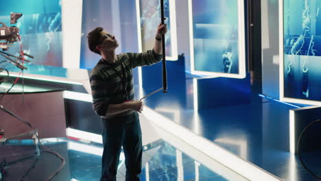 technician in broadcast studio adjusting boom pole, preparing for filming. modern media production environment with equipment, lighting, and professional studio setup visible in background