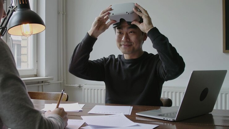 Un uomo con un maglione scuro prende un visore VR bianco e lo testa in ufficio tra laptop e documenti