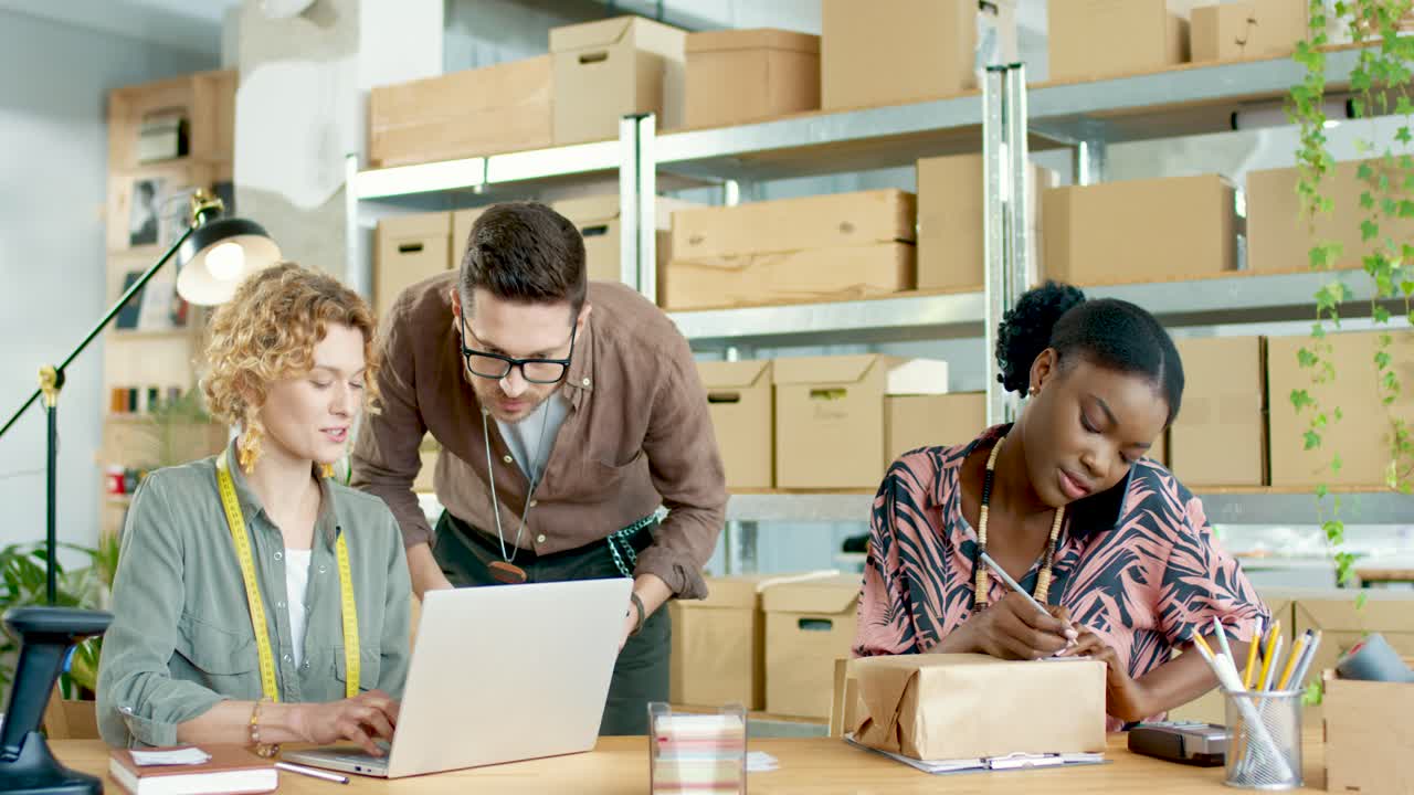 Multiethnic designers team typing on tablet, browsing on laptop and talking on the phone in fashion clothing shop