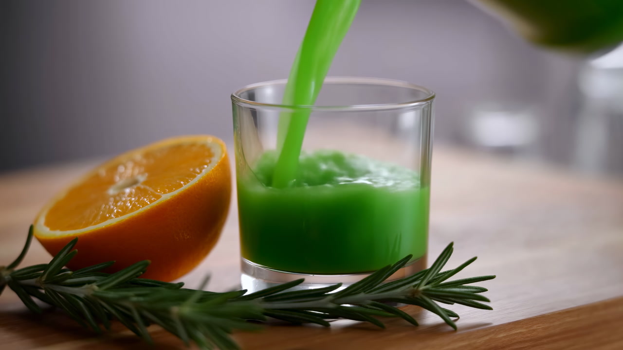 Green Juice Pouring Next To Orange And Rosemary On Wooden Board, Closeup Slowmotion Pour Into Clear Glass,