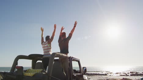 happy caucasian gay male couple standing in car raising arms and holding hands on sunny day at beach