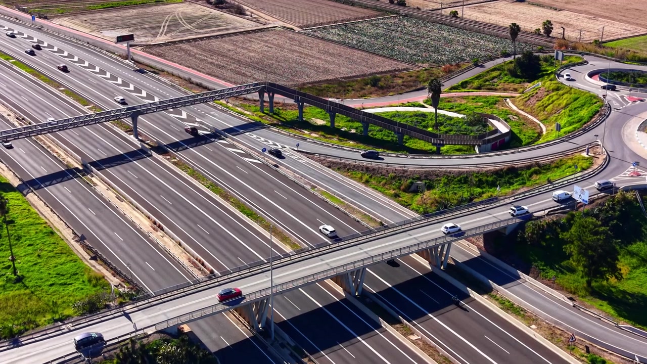 Aerial view of overlapping highway bridges and roundabout, Spain. Two overpasses cross a wide motorway near farmland and a circular junction.