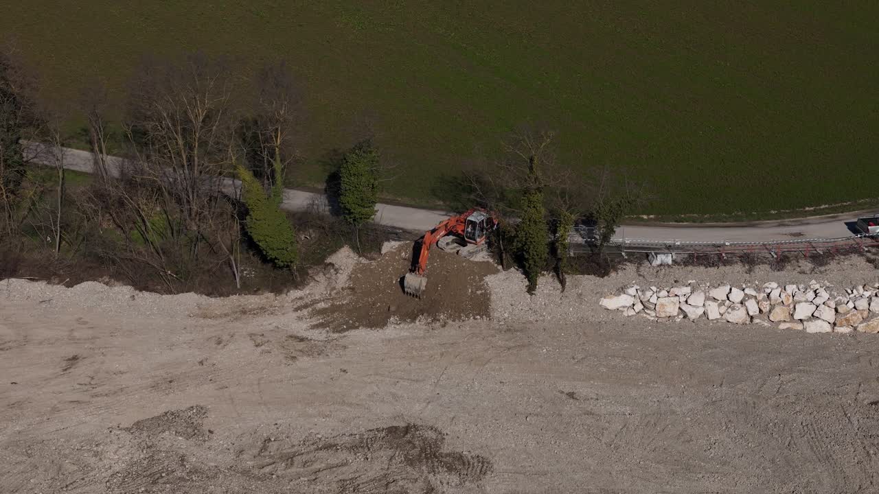 Un escavatore e un camion sono visibili in un cantiere con un grande cumulo di rocce e un campo verde sullo sfondo