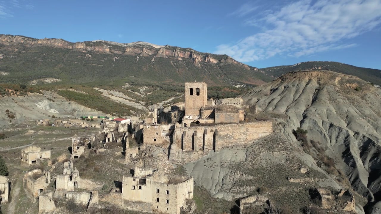 Vista aerea che sorvola la desolata e abbandonata città fantasma di Esco, rivelando i suoi edifici in pietra in rovina e l'antica chiesa tra le brulle colline dell'Aragona, Spagna.