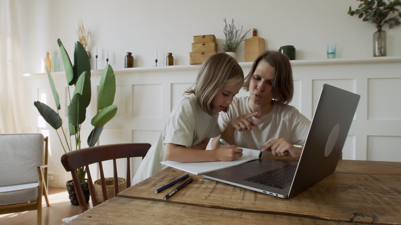 una linda chica rubia hace su tarea con la ayuda de su madre mientras mira la pantalla del portátil