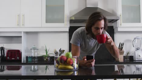Hombre-Caucásico-Tomando-Café-Y-Usando-Un-Teléfono-Inteligente-En-La-Cocina-De-Casa