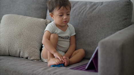 Cute-young-latin-toddler-kid-baby-boy-in-a-light-blue-baby-romper-sitting-on-a-grey-couch-watching-cartoons-on-a-purple-tablet