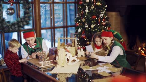 two elves are sitting at the table with the children and show them letters of congratulations and new year's gifts against the background of the christmas tree and the fireplace near the window in the house