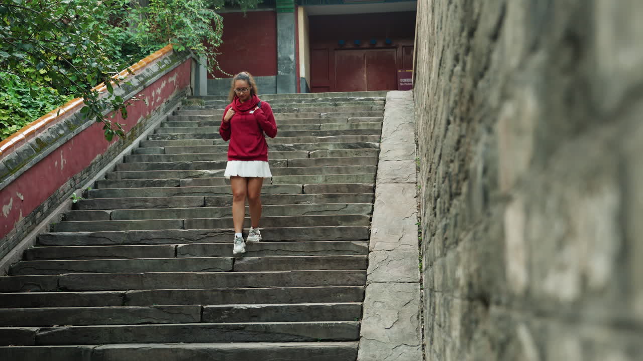 Người phụ nữ châu Á bước lên các bậc thang đền, mặc áo khoác đỏ, váy trắng và giày thể thao, leo lên cầu thang đá hai bên là bức tường phong hóa và cây xanh tươi tốt, một du khách bình thường đang chỉnh lại mũ áo khoác, vẻ mặt trầm tư, yên tĩnh.