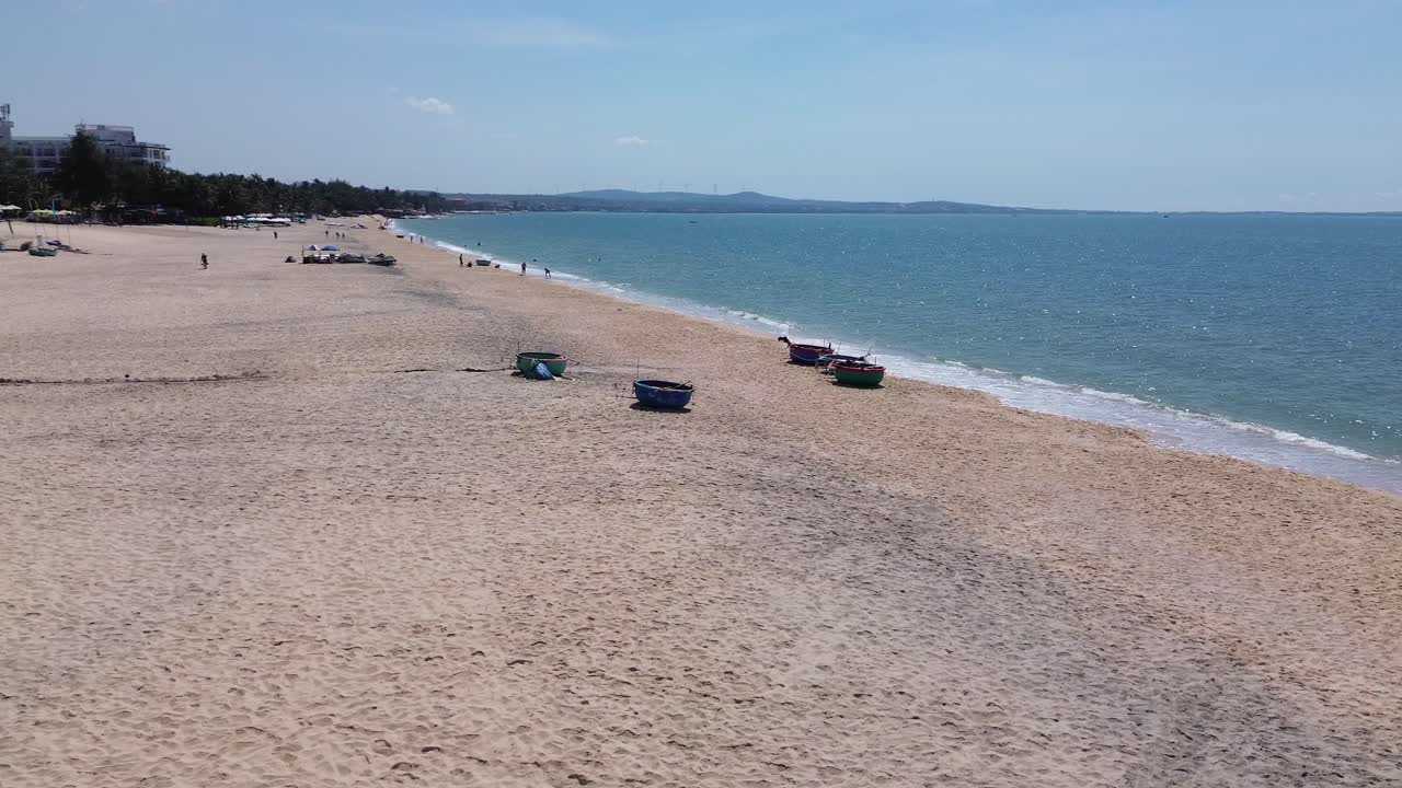Drone flies in a half-circle from right to left, keeping Mui Ne beach centered with fishing buckets on sand, revealing resorts, white shore, and the sea horizon.