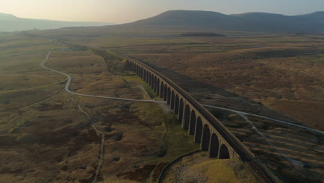 Hohe-Drohnenaufnahme-Aus-Der-Luft,-Die-Sich-Bei-Atemberaubendem-Sonnenaufgang-Im-Sommer-In-Den-Yorkshire-Dales,-England,-Großbritannien,-Mit-Ingleborough-Im-Hintergrund-Um-Die-Ribblehead-Viadukt-Zugbrücke-Dreht