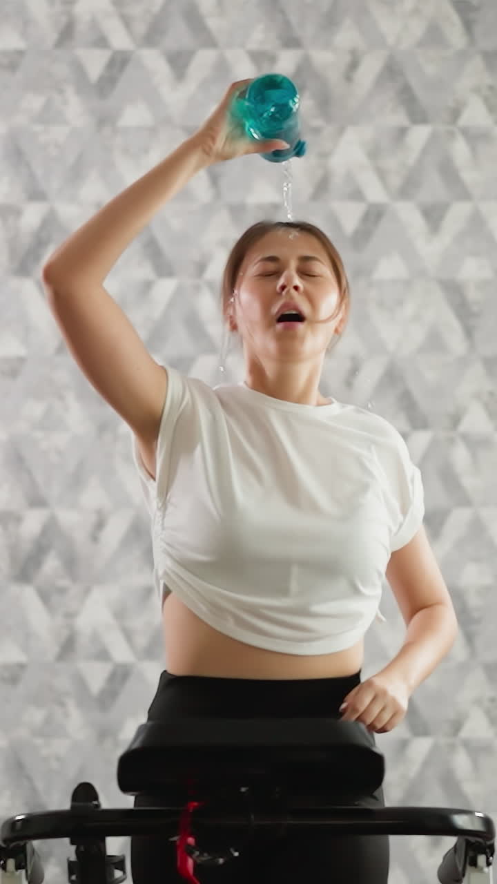 Woman pours water from bottle on head running at home. Tired lady cools down exercising on treadmill in domestic gym. Sportswoman deals with overheat at training