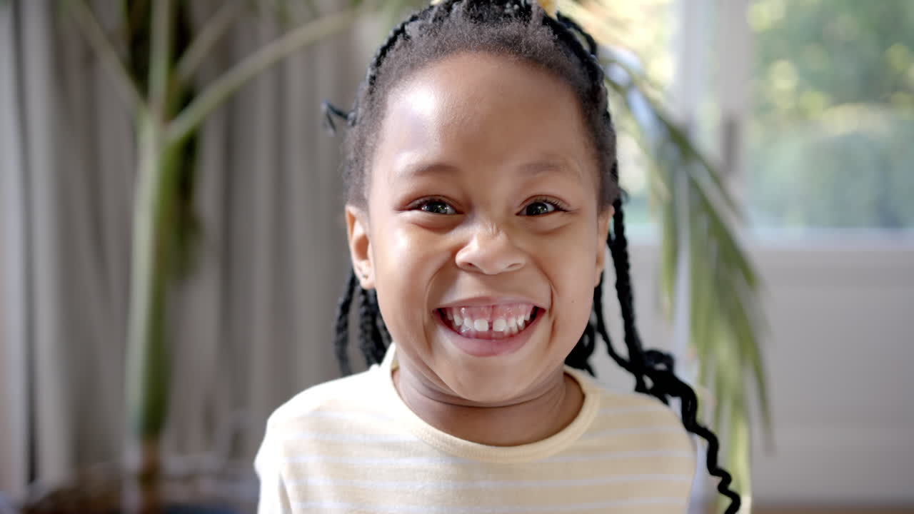 Retrato de menina afro-americana feliz com cabelo trançado sorrindo perto de uma janela ensolarada, em câmera lenta