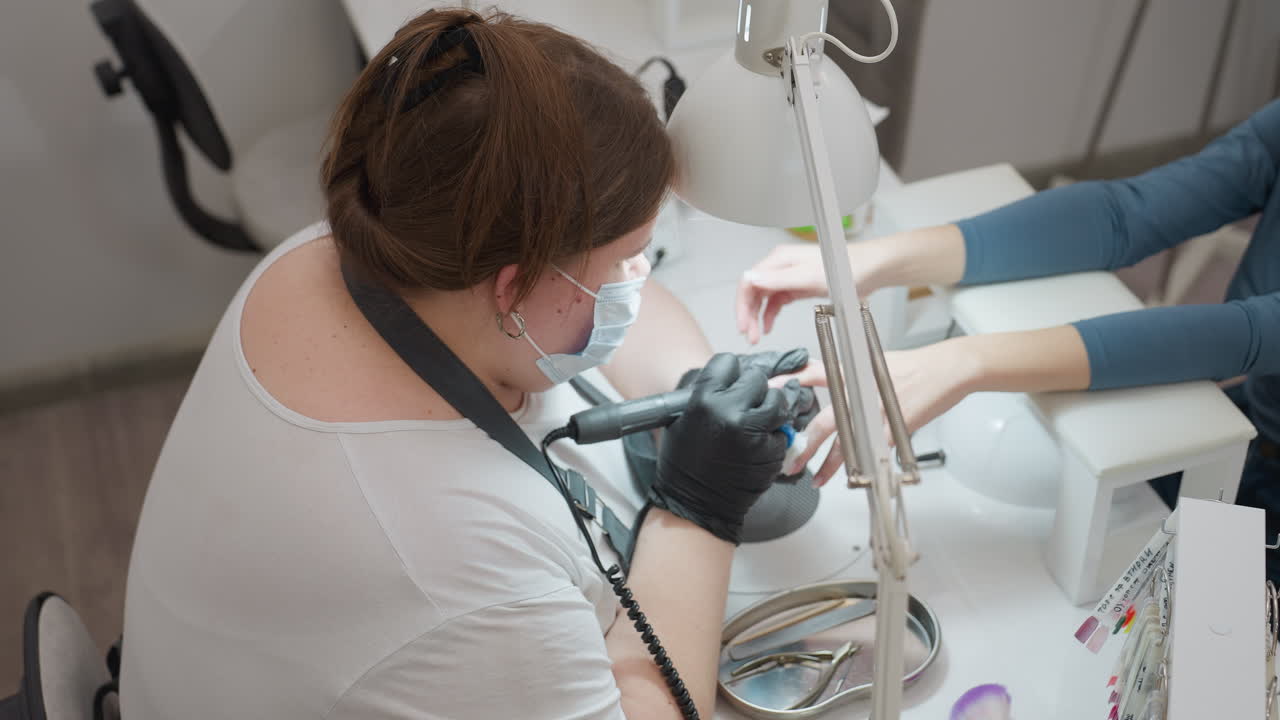 Top view of nail technician with tied blond hair, wearing face mask and apron, carefully filing and brushing customer nails with precision under white salon desk lamp in clean professional environment