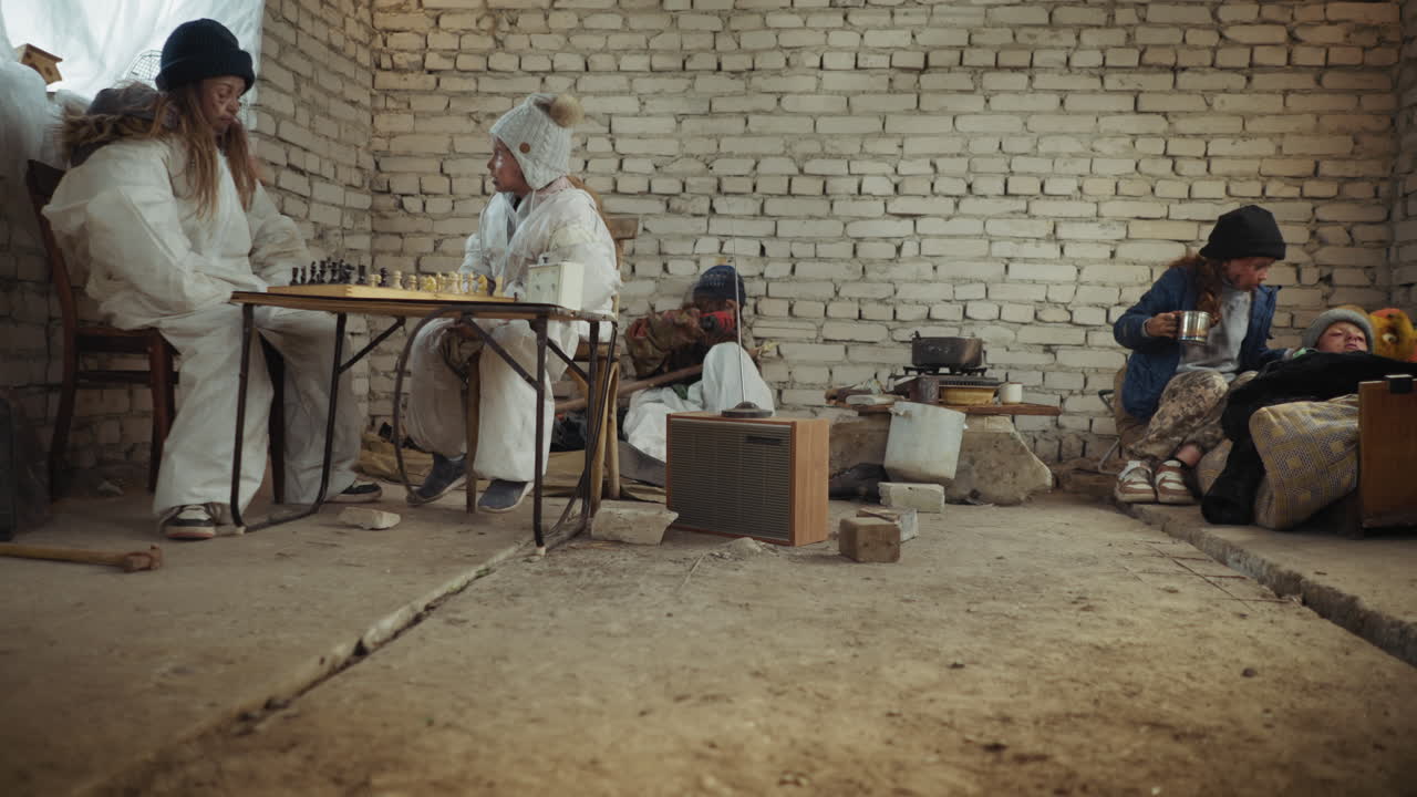 Бездомные дети в холодной заброшенной комнате: двое напряженно играют в шахматы за столом, один точит дерево, чтобы выжить, другой ухаживает за ребенком на самодельной кровати возле кухонной утвари и обогревателя, создавая суровую картину лишений