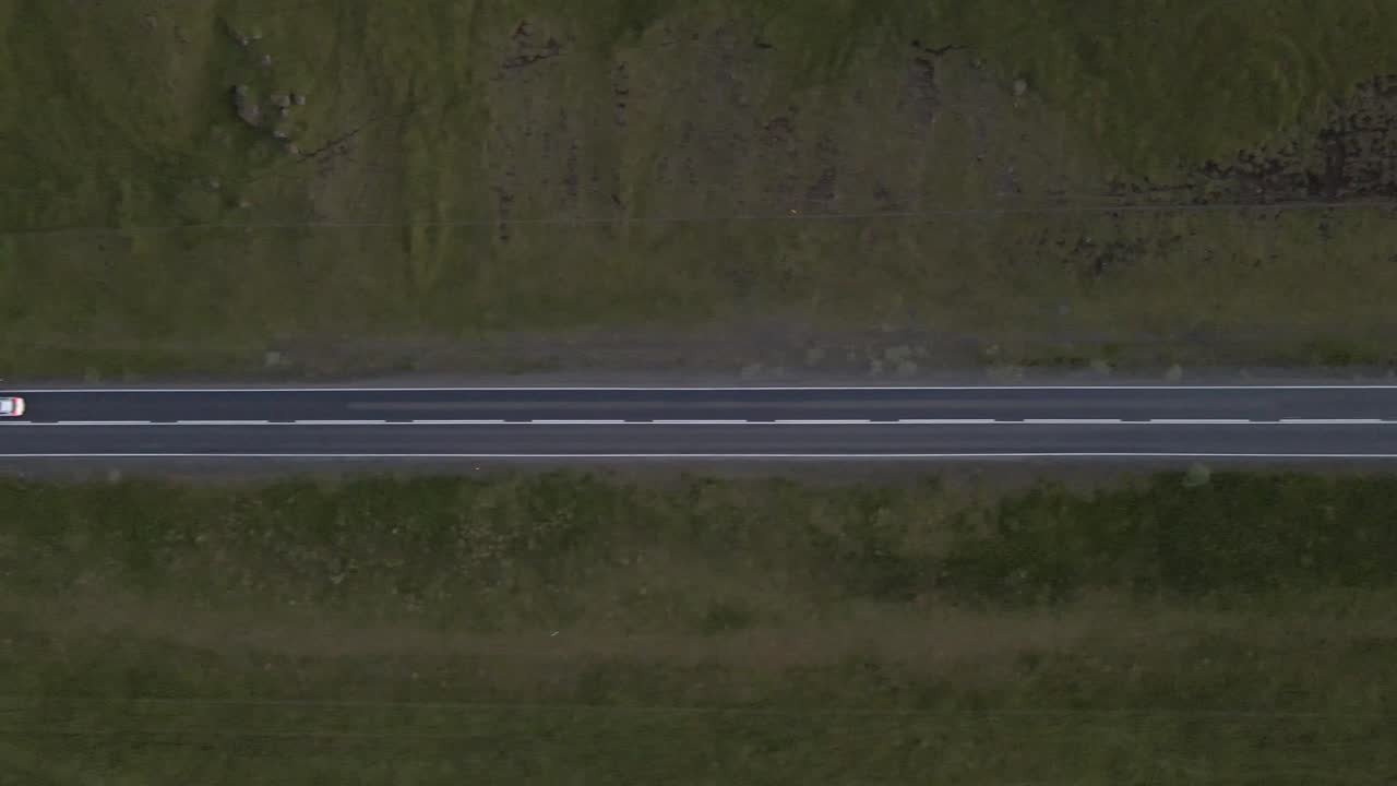 Night descends as headlights carve a path through the darkness, an overhead shot following a lone car's journey down a straight, secluded road