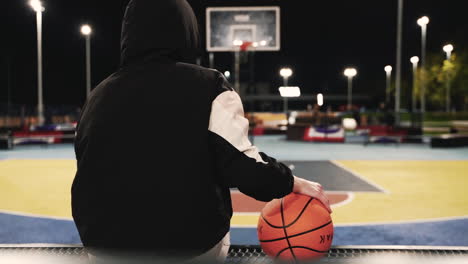 Rückansicht-Einer-Basketballspielerin-Im-Hoodie,-Die-Sitzt-Und-Den-Ball-Hält,-Während-Sie-Nach-Ihrer-Trainingseinheit-Nachts-Auf-Dem-Außenplatz-Eine-Pause-Macht