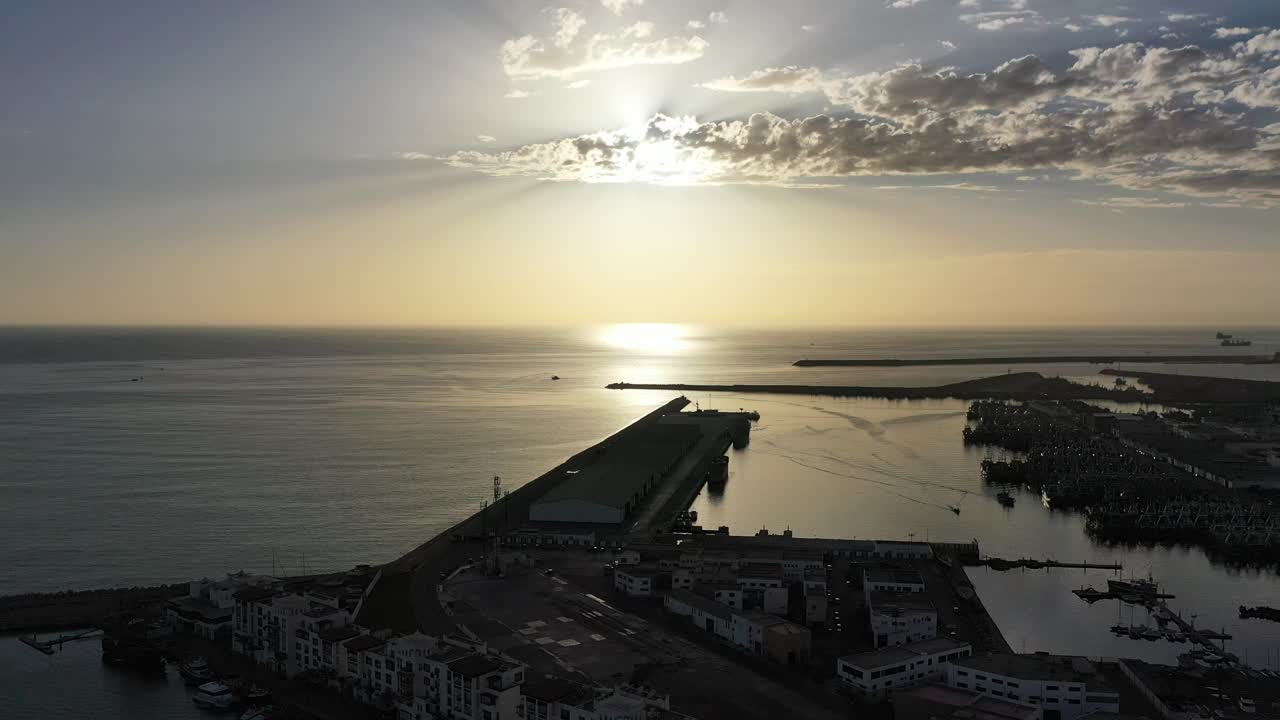 Riprese mozzafiato con drone che catturano l'ora d'oro ad Agadir. Mostra il sole che tramonta sul mare calmo, le silhouette del porto industriale e turistico, e uno splendido cielo nuvoloso in Marocco
