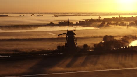 low aerial shot of a dramatic silhouetted windmill and farm fields with a light fog clinging to the ground in early morning bright orange sun