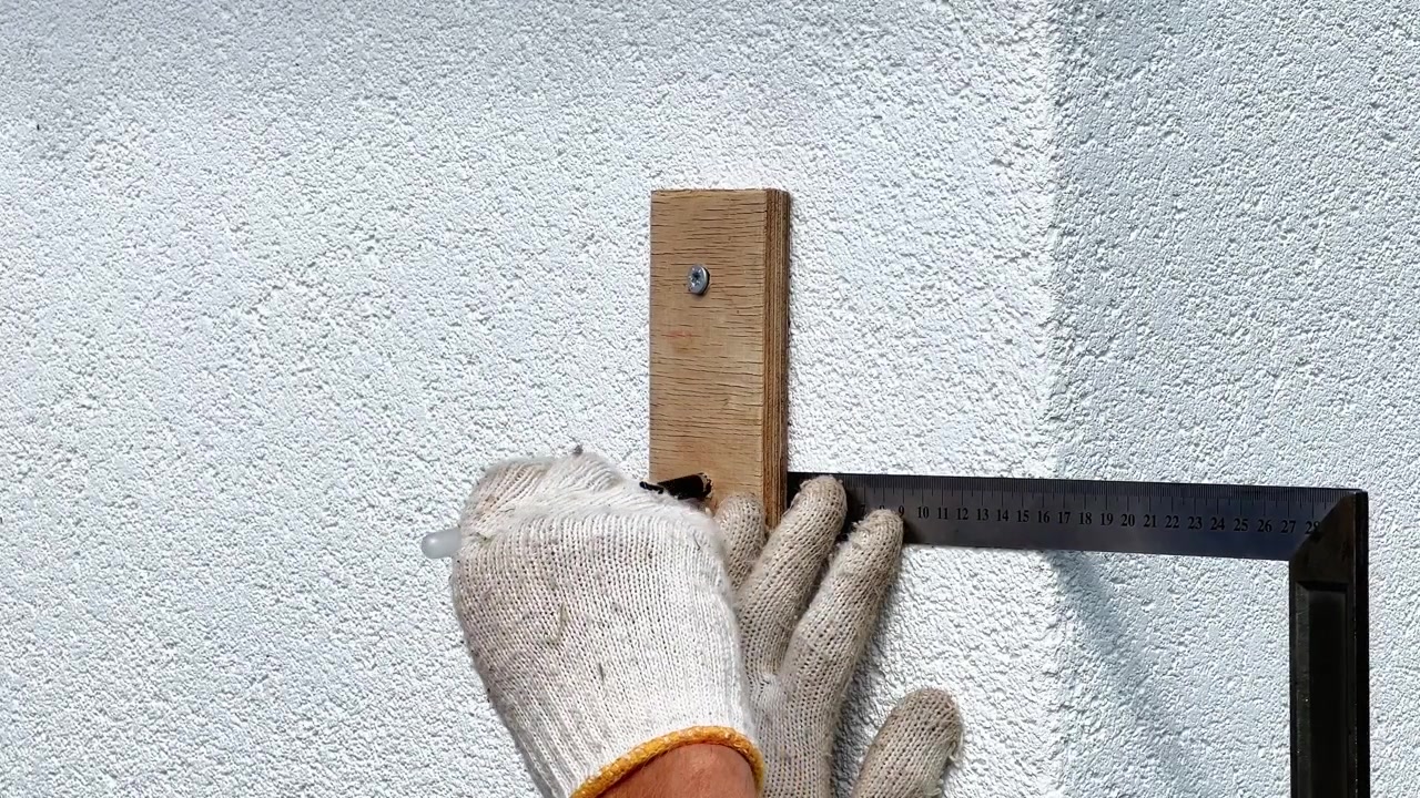 Close-up shot of a gloved worker using a steel try square ruler and pencil to measure alignment on a white wall. Ideal for construction tutorials and DIY guides.