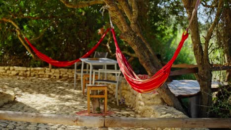 Beautiful-red-hammocks-hanging-against-some-trees-with-outdoor-table-and-chairs,-relax-in-nature