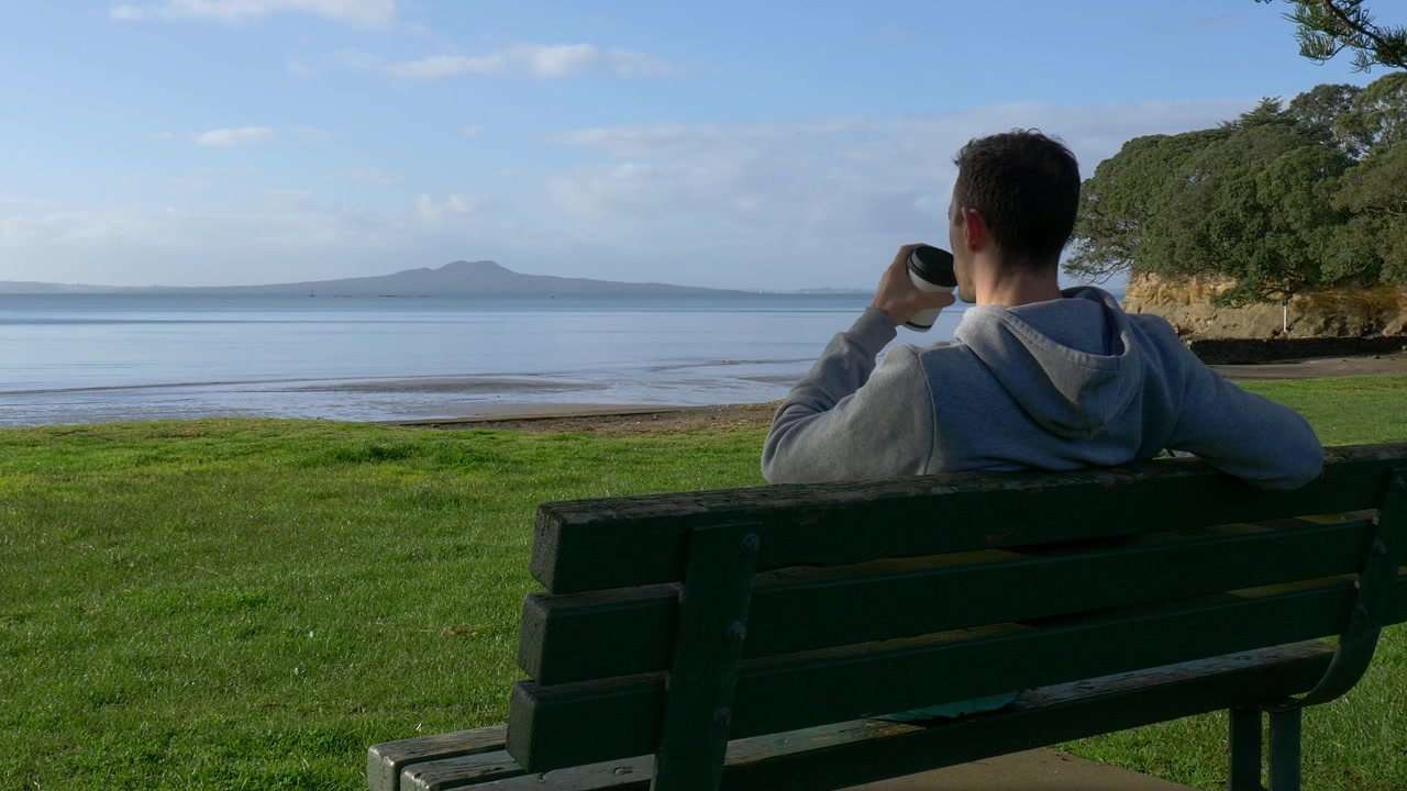 Young caucasian man drinking coffee sitting on a bench at the beach looking at Rangitoto island in Auckland, New Zealand