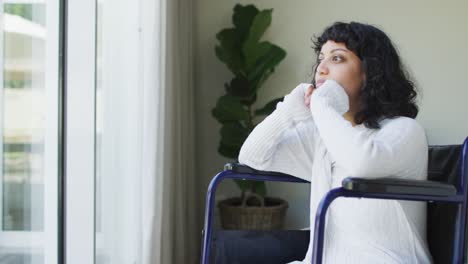 depressed biracial disabled woman in wheelchair looking out of window, then covering eyes