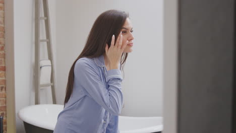 woman wearing pyjamas at home in modern bathroom using moisturiser looking in mirror