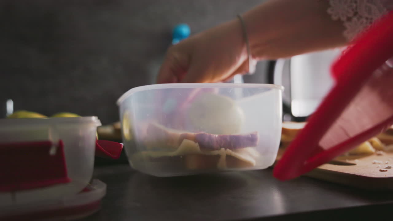 closeup view of hand placing cheese sandwich slices into clear plastic container on kitchen countertop next to sliced bread and green apples in bright morning light preparing healthy school lunch