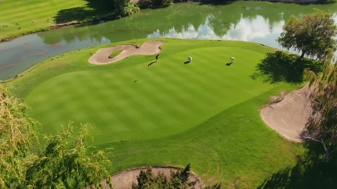 Tomada de drone com aproximação de um green de ilha rodeado por águas calmas e árvores altas, com golfistas visíveis na superfície de tacada.
