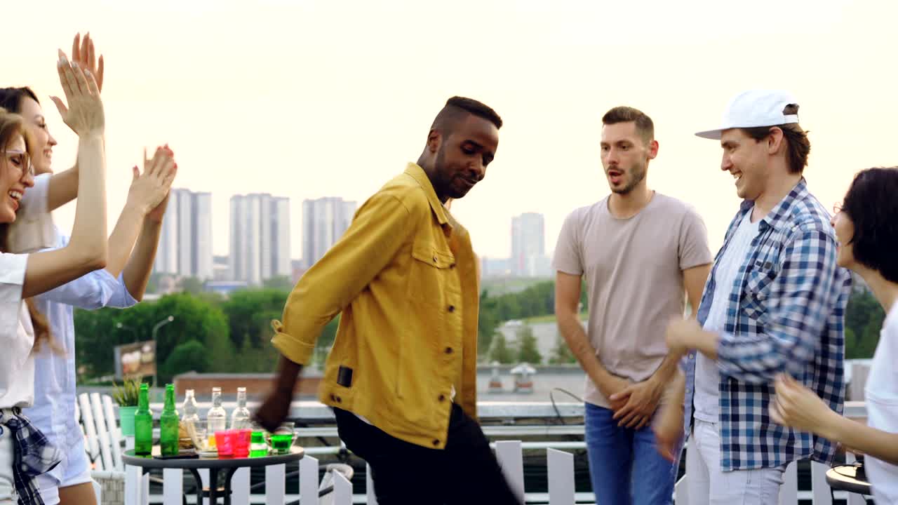 Joyful African American man in trendy clothes is dancing with friends multiracial group having fun at party on roof on summer day. Leisure, joy and friendship concept.