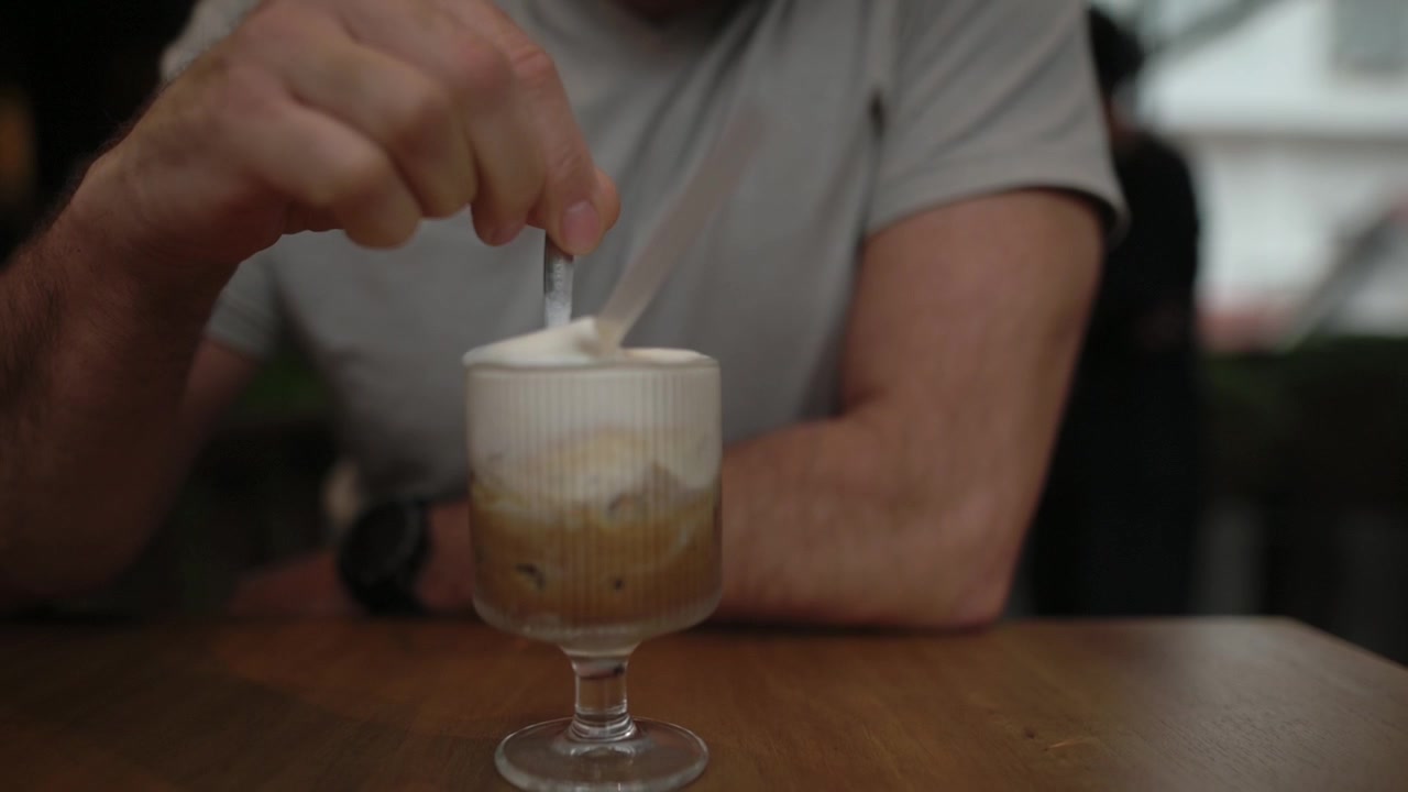 Una mano mescola caffè vietnamita al cocco in un bicchiere con un cucchiaio, mentre una spessa schiuma di cocco si avvolge nello strato di espresso. Si intravedono polso e braccio in questa scena casuale da caffè al chiuso, caratterizzata da un movimento intimo e un'atmosfera calda.