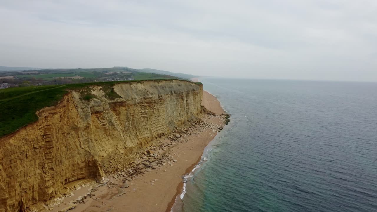 Jurassic Coast cliff with a golden beach and blue see beneath. Aerial footage moves backwards and raises up to show this grass top cliff and surrounding area on a spring cloudy day in Dorset.