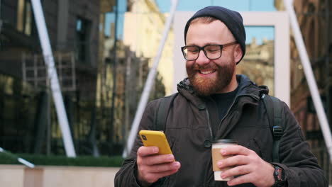 smiling man with beard and glasses using a smartphone and holding a coffee cup in a modern city setting