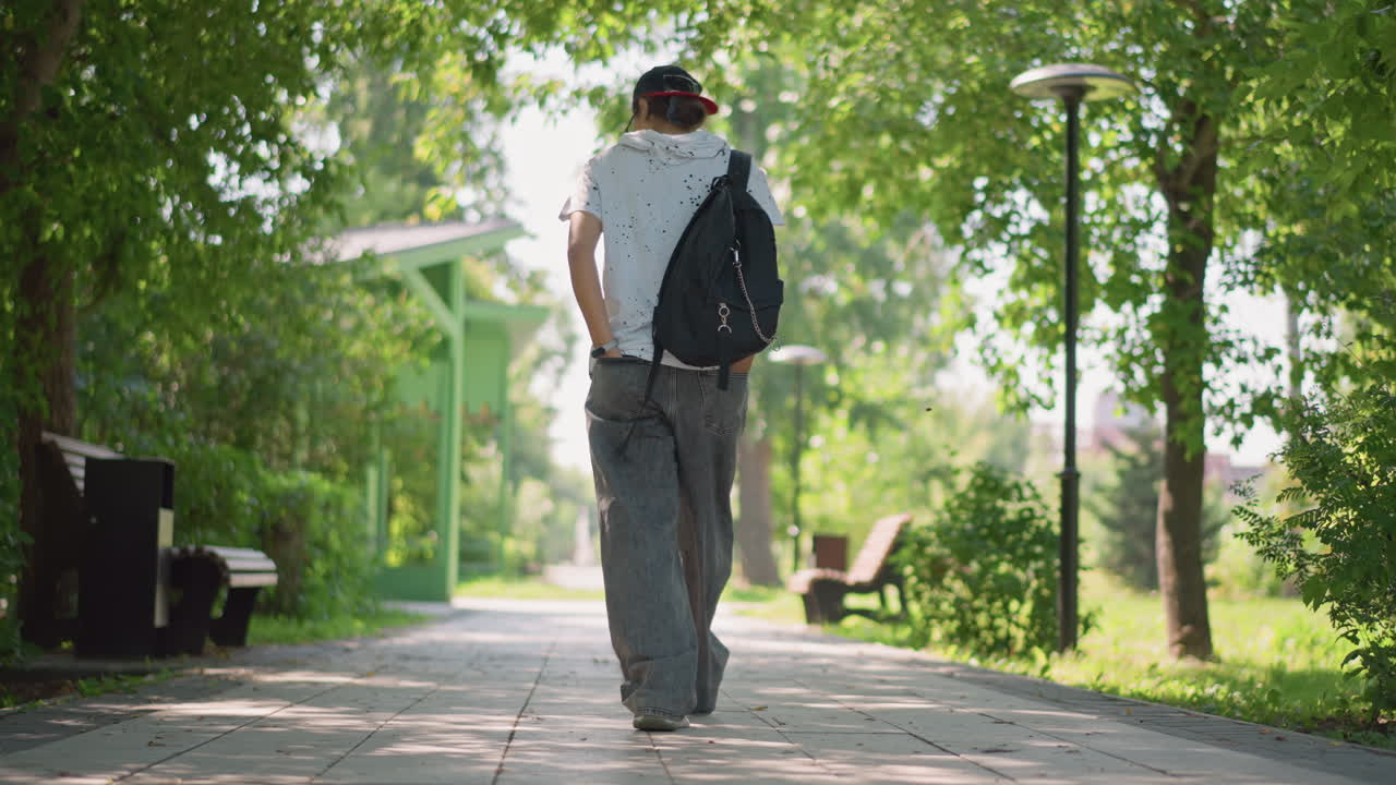 Person progressing through city park walkway, Male individual moving along shaded path with backpack, Man with cap and backpack navigating treecovered walkway during morning journey