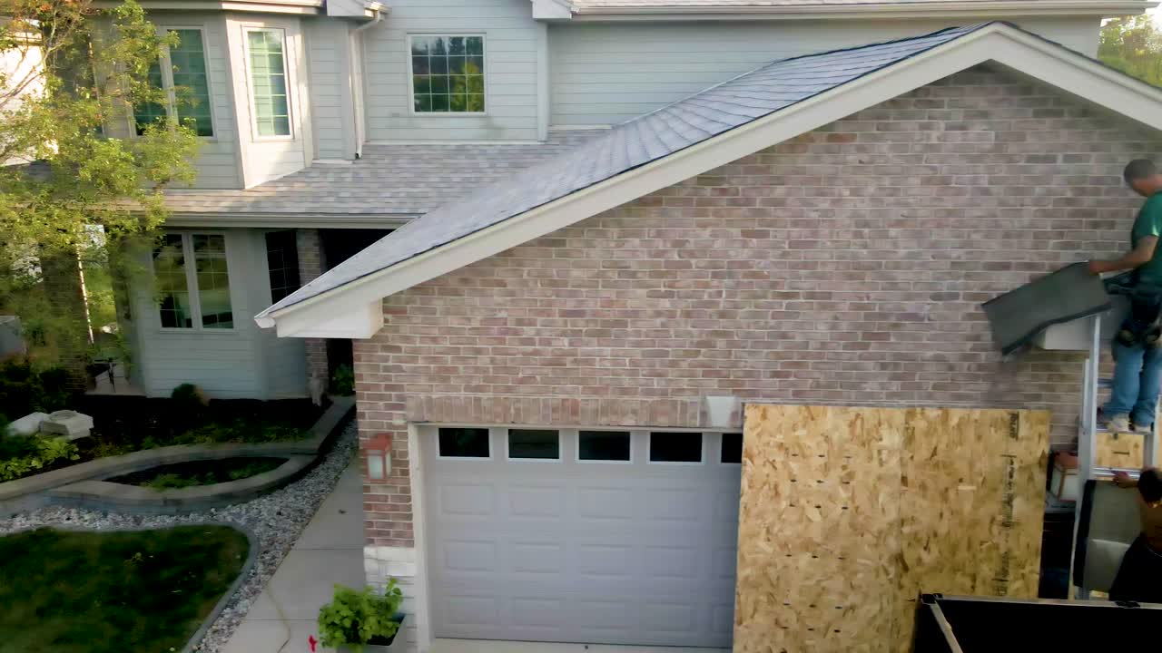 Construction worker repairing the exterior of a brick house, standing on a ladder and working on the roof area above the garage."
