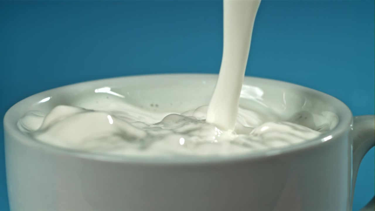 青い背景で白いマグカップに牛乳をスローモーションで注ぐ。