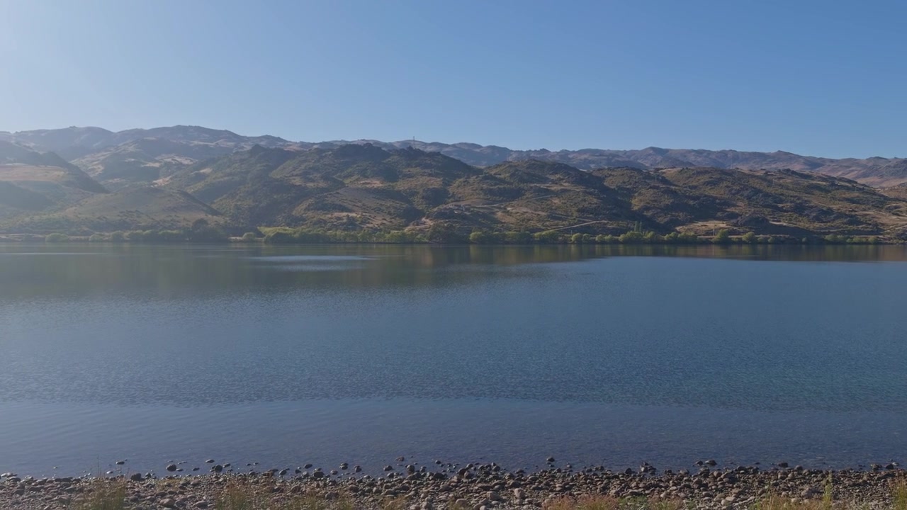 Smukt panoramaudsigt over Central Otago med roligt, reflekterende vand, bølgende bjerge og grønne træer under en klar blå himmel.