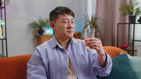 Thirsty-asian-man-sitting-at-home-holding-glass-of-natural-aqua-make-sips-drinking-still-water